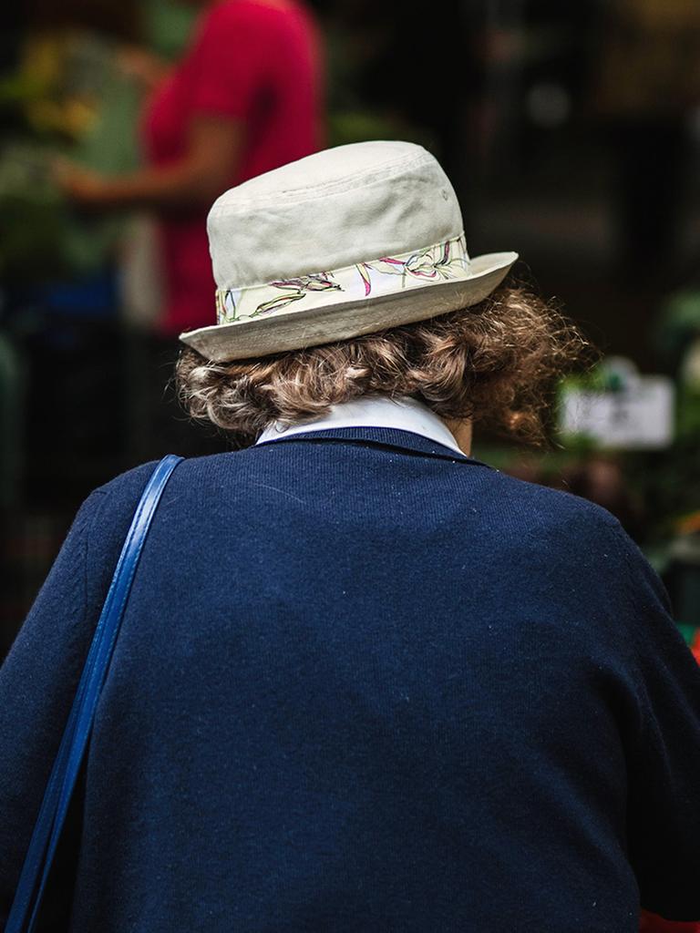 Eine ältere Frau mit Hut und dem Rücken zur Kamera geht über einen Gemüsemarkt. 