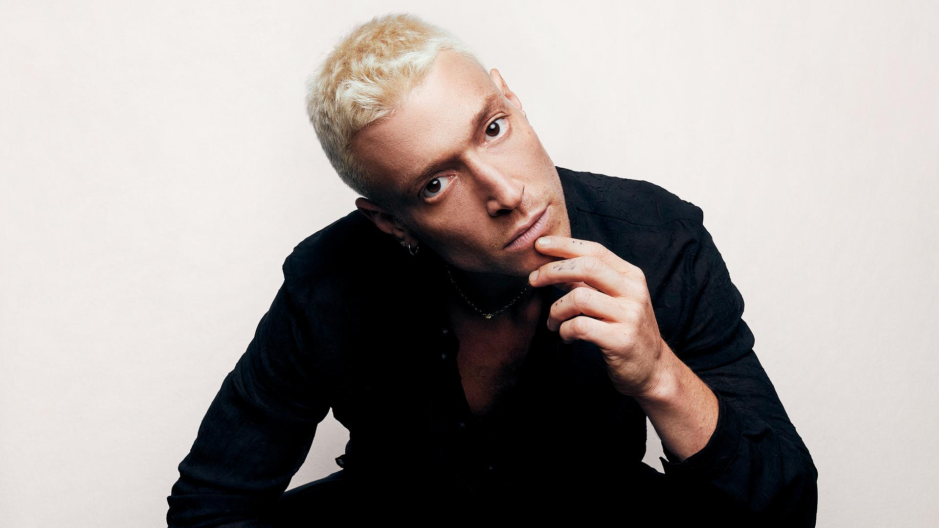 Portrait eines jungen Mannes, mit kurzen blondierten Haaren, der die Finger seiner linken Hand am Kinn hält und den Betrachter anblickt. Portrait eines jungen Mannes, mit kurzen blondierten Haaren, der die Finger seiner linken Hand am Kinn hält und den Betrachter anblickt.