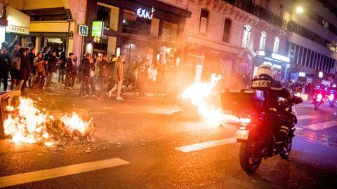 Frankreich, Paris: Ein Feuer brennt auf einer Straße während Menschen nach der Verabschiedung einer umstrittenen Rentenreform protestieren und die Polizei im Einsatz ist.