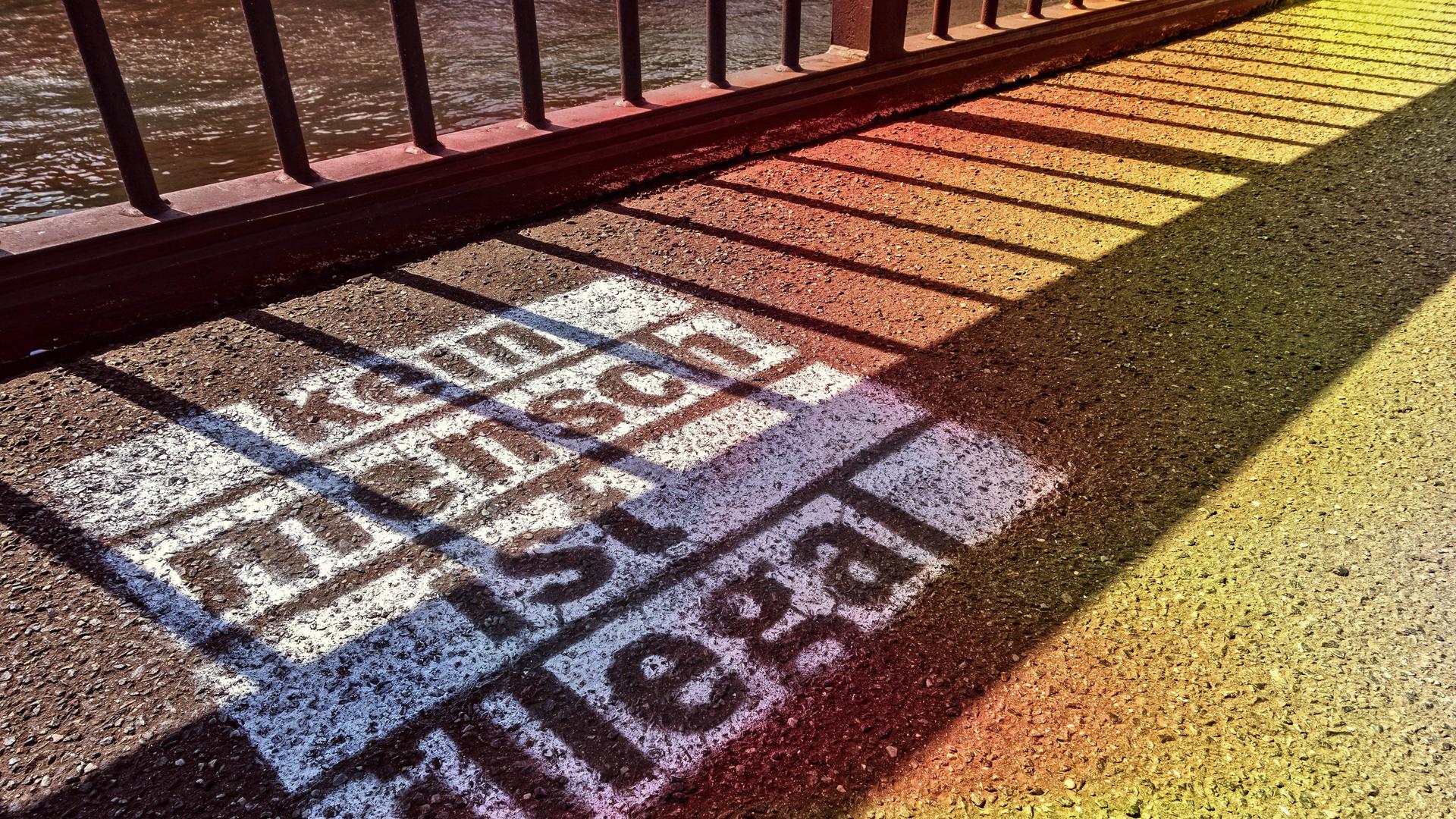 Auf den Asphalt vor einem Zaun ist mit weißer Farbe der Schriftzug gesprüht: "Kein Mensch ist illegal". Auf den Asphalt vor einem Zaun ist mit weißer Farbe der Schriftzug gesprüht: "Kein Mensch ist illegal".