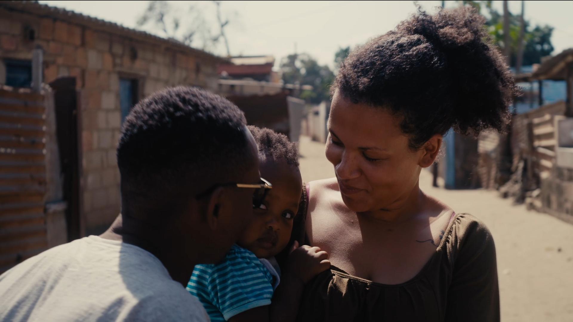 Filmszene aus dem Dokumentarfilm "The Homes We Carry". Zu sehen ist eine junge mosambikanische Frau (vermutlich die Protagonistin des Film) mit Kind auf dem Arm sowie die Rückansicht eines Mannes. Filmszene aus dem Dokumentarfilm "The Homes We Carry". Zu sehen ist eine junge mosambikanische Frau (vermutlich die Protagonistin des Film) mit Kind auf dem Arm sowie die Rückansicht eines Mannes.