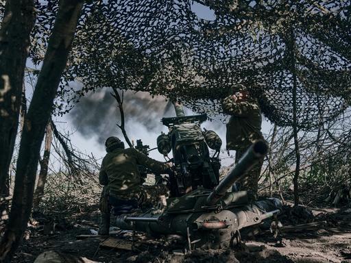Zwei Soldaten stehen an einer Kanone. Sie stehen in einem Gebüsch, das mit Tarnnetz umhüllt ist.