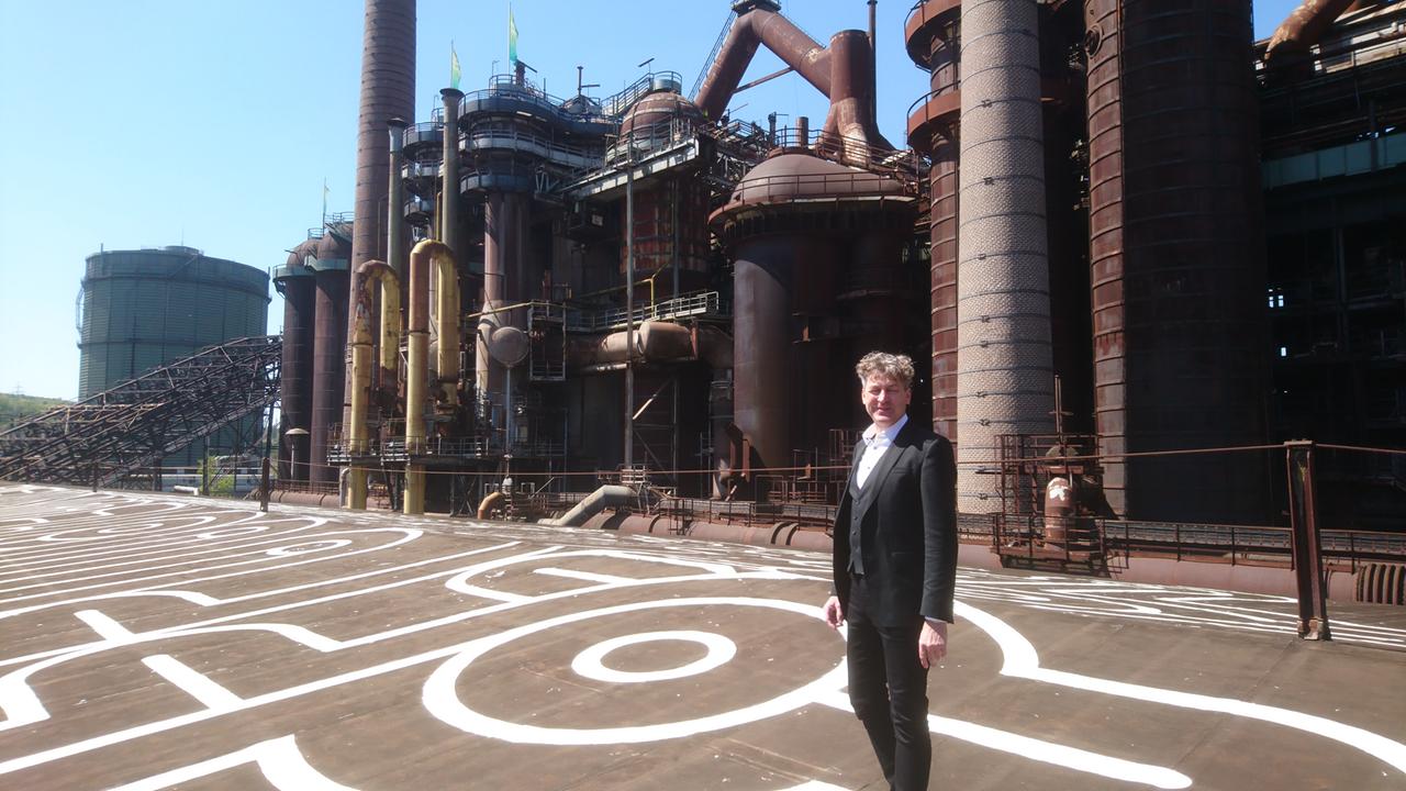 Ralf Beil, der Generaldirektor des Weltkulturerbes Völklinger Hütte, auf dem Dach der Möllerhalle. Hinter ihm ist ein alter Industriekomplex aus Hochöfen zu sehen. Ralf Beil, der Generaldirektor des Weltkulturerbes Völklinger Hütte, auf dem Dach der Möllerhalle. Hinter ihm ist ein alter Industriekomplex aus Hochöfen zu sehen.