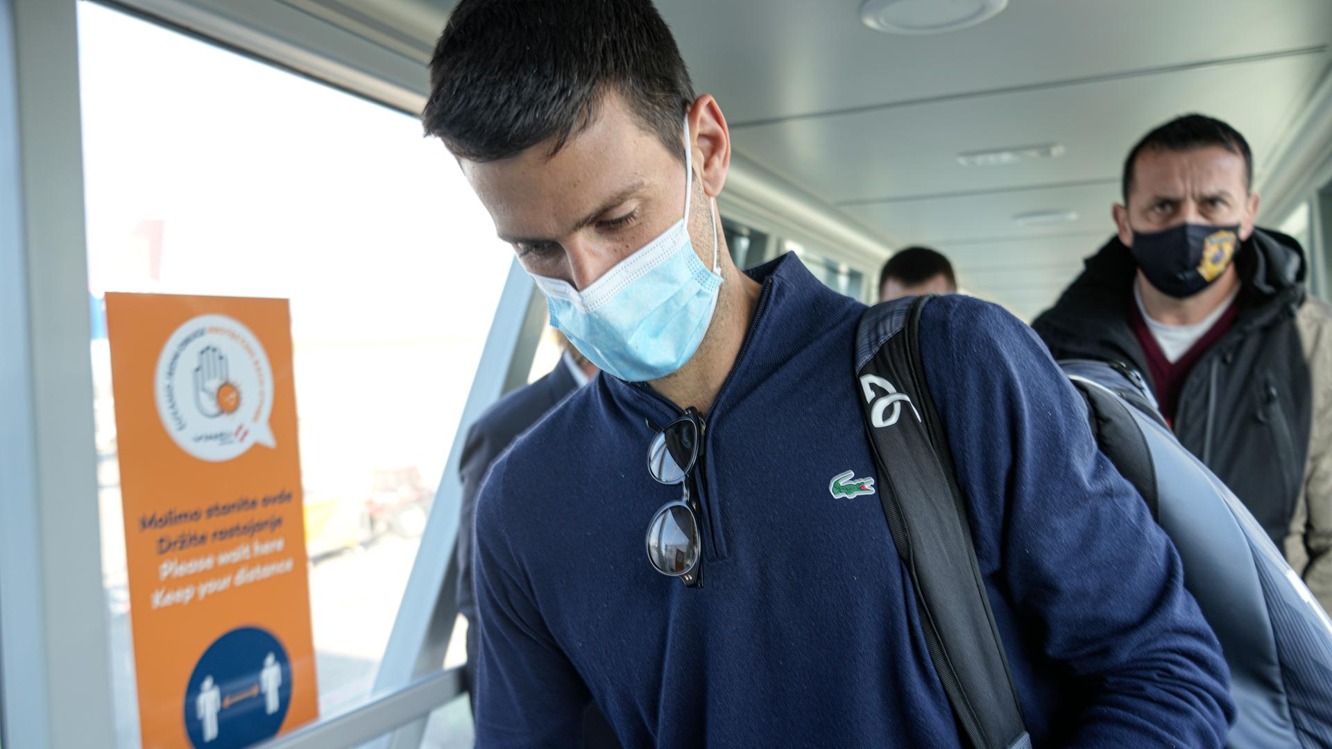 Djokovic geht mit Maske einen Gang entlang und schaut nach unten. Dahinter zwei weitere Männer. Djokovic geht mit Maske einen Gang entlang und schaut nach unten. Dahinter zwei weitere Männer.