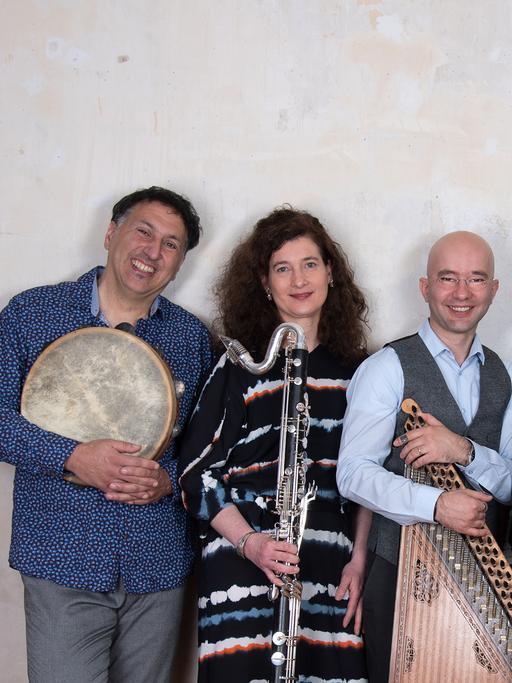 Fünf Menschen stehen nebeneinander vor einer grauen Wand und lächeln. Sie tragen Instrumente in den Händen: Trommeln, Akkordeon und eine Klarinette. Zusammen bilden sie das Ensemble FisFüz.