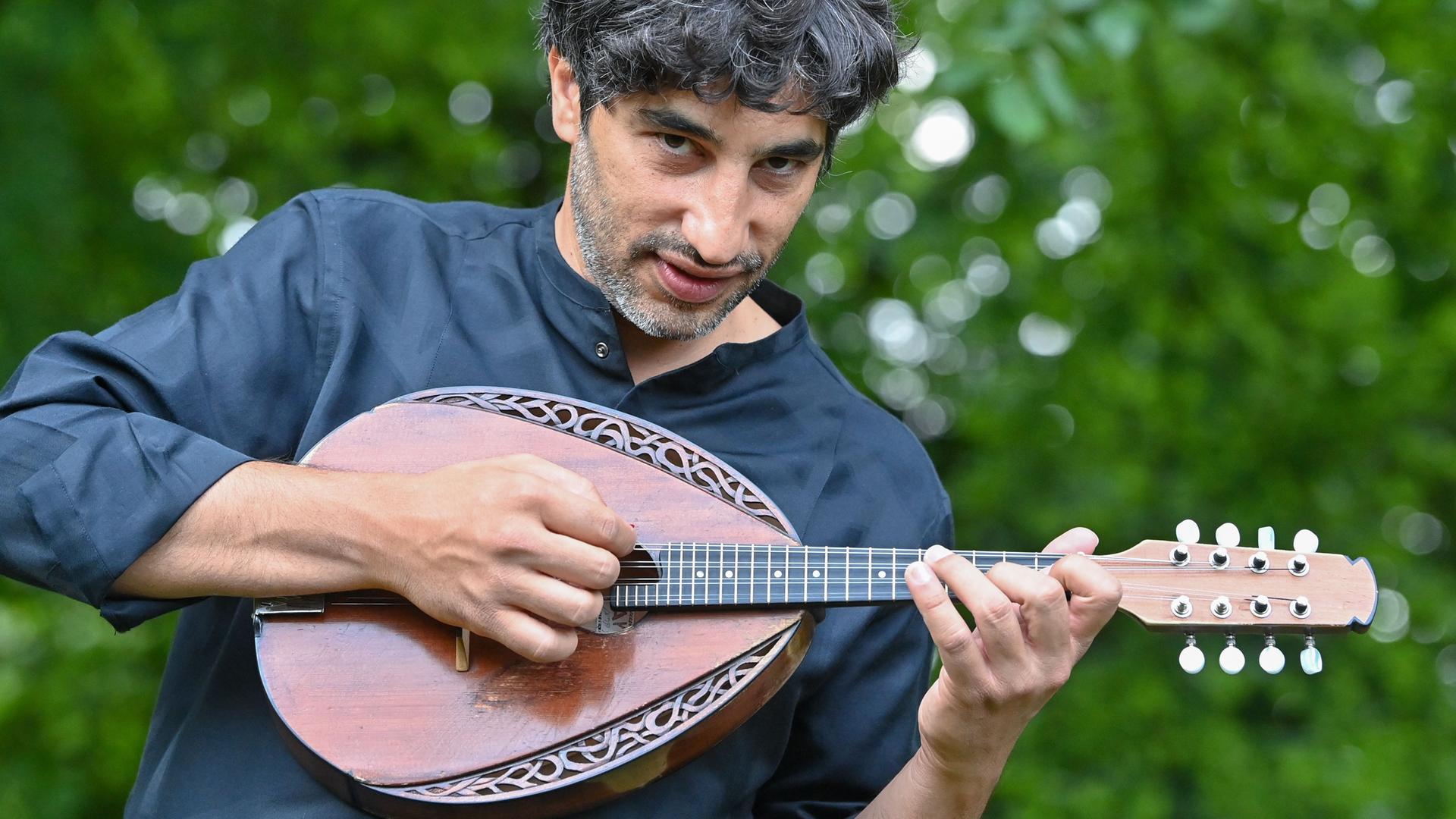 Avi Avital steht mit einer Mandoline im Freien vor Grünpflanzen. Er hat lockige, dunkle Haare, trägt ein dunkelfarbiges Hemd und hält eine Mandoline vor der Brust.