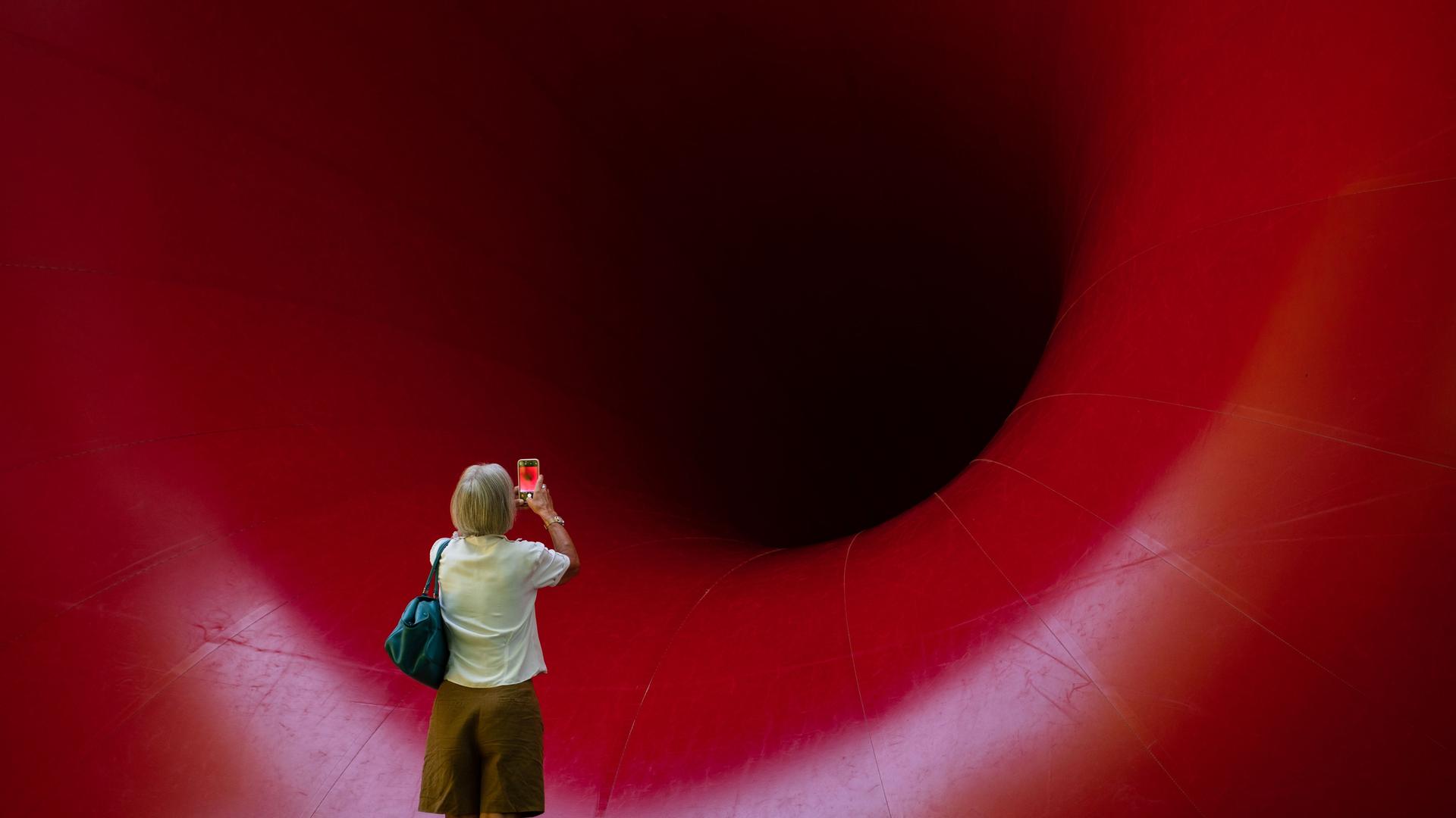 Eine Besucherin steht vor einer riesigen Großskulptur von Anish Kapoor im Skulpturenpark Waldfrieden. Dabei steht sie winzig wirkend vor einem riesigen roten Trichter. Eine Besucherin steht vor einer riesigen Großskulptur von Anish Kapoor im Skulpturenpark Waldfrieden. Dabei steht sie winzig wirkend vor einem riesigen roten Trichter.