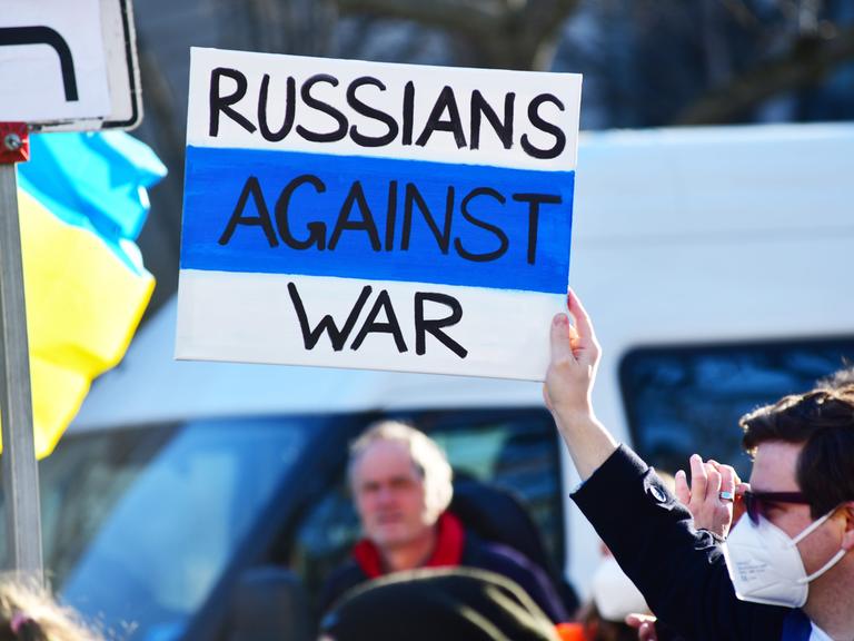 Demonstration in Berlin gegen den russischen Krieg in der Ukraine. Links sieht man eine blau-gelbe Fahne, rechts hält ein Mann ein Plakat mit der Aufschrift "Russians against War" in die Luft.