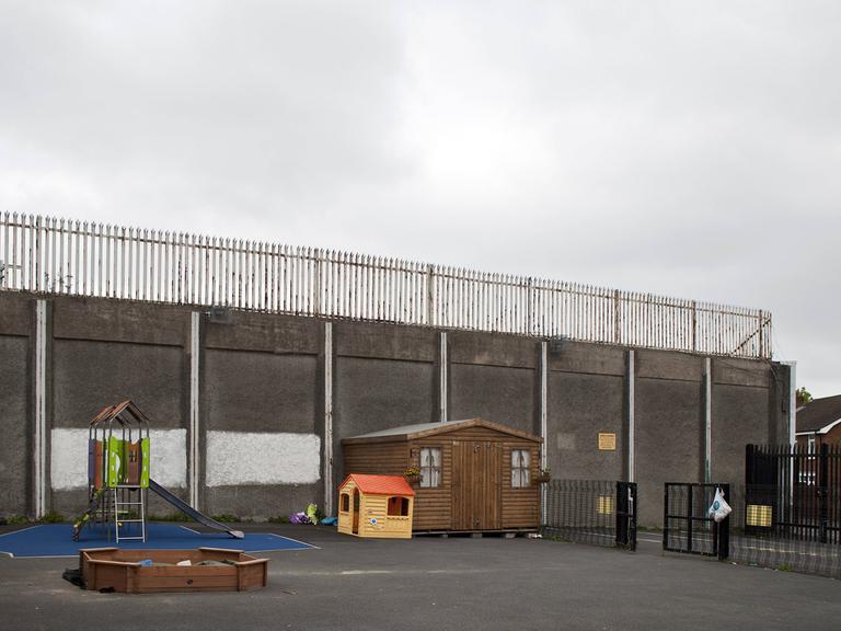 Ein Spielplatz in Belfast, Nordirland, vor einer hohen Mauer, die katholische und protestantische Wohngebiete trennt.