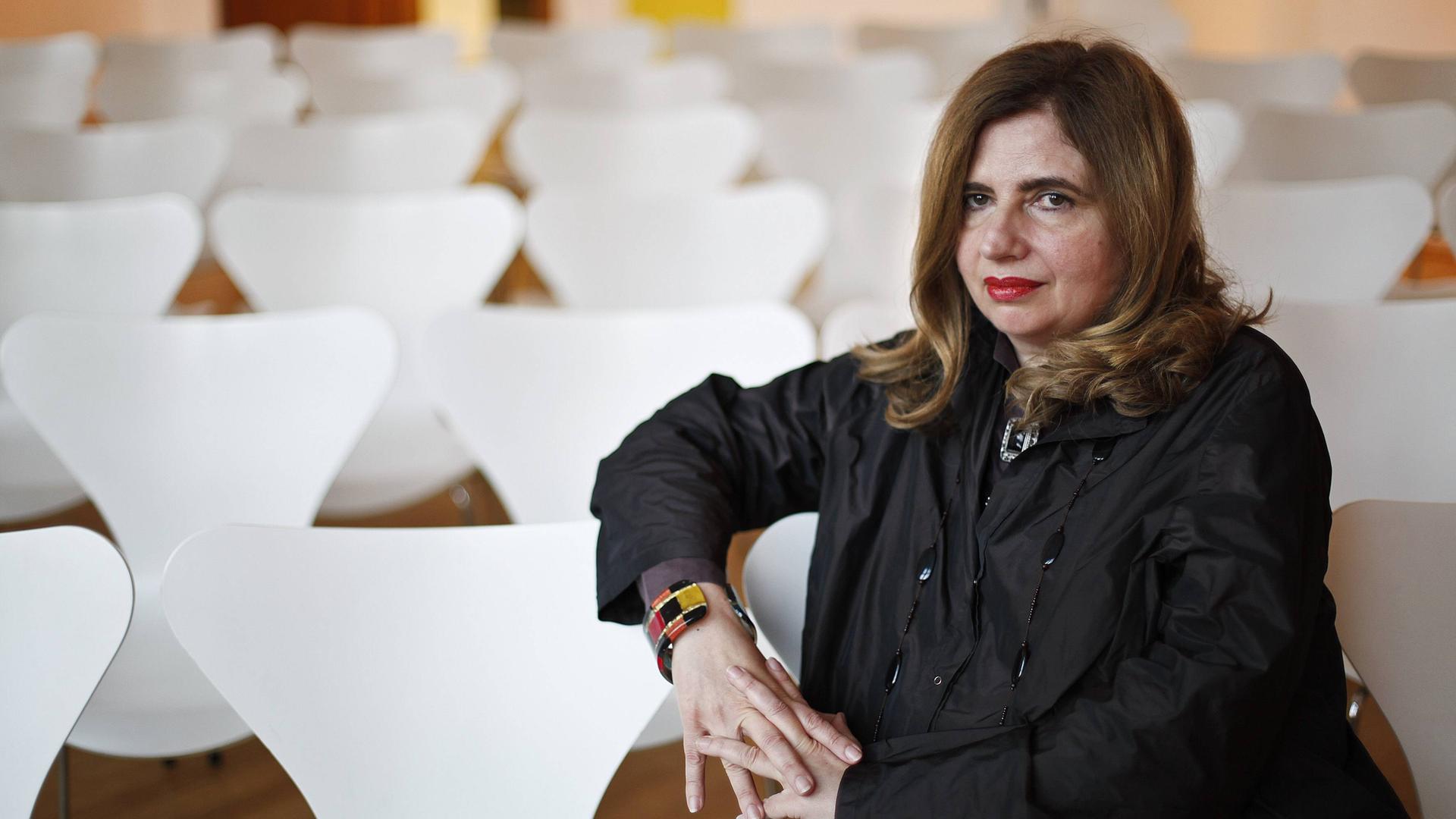 Sibylle Lewitscharoff sitzt in Porträtpose auf einem weißen Stuhl und hat den Arm auf die Lehne gelegt. Neben und hinter ihr sind weitere Stühle zu sehen. Sibylle Lewitscharoff sitzt in Porträtpose auf einem weißen Stuhl und hat den Arm auf die Lehne gelegt. Neben und hinter ihr sind weitere Stühle zu sehen.