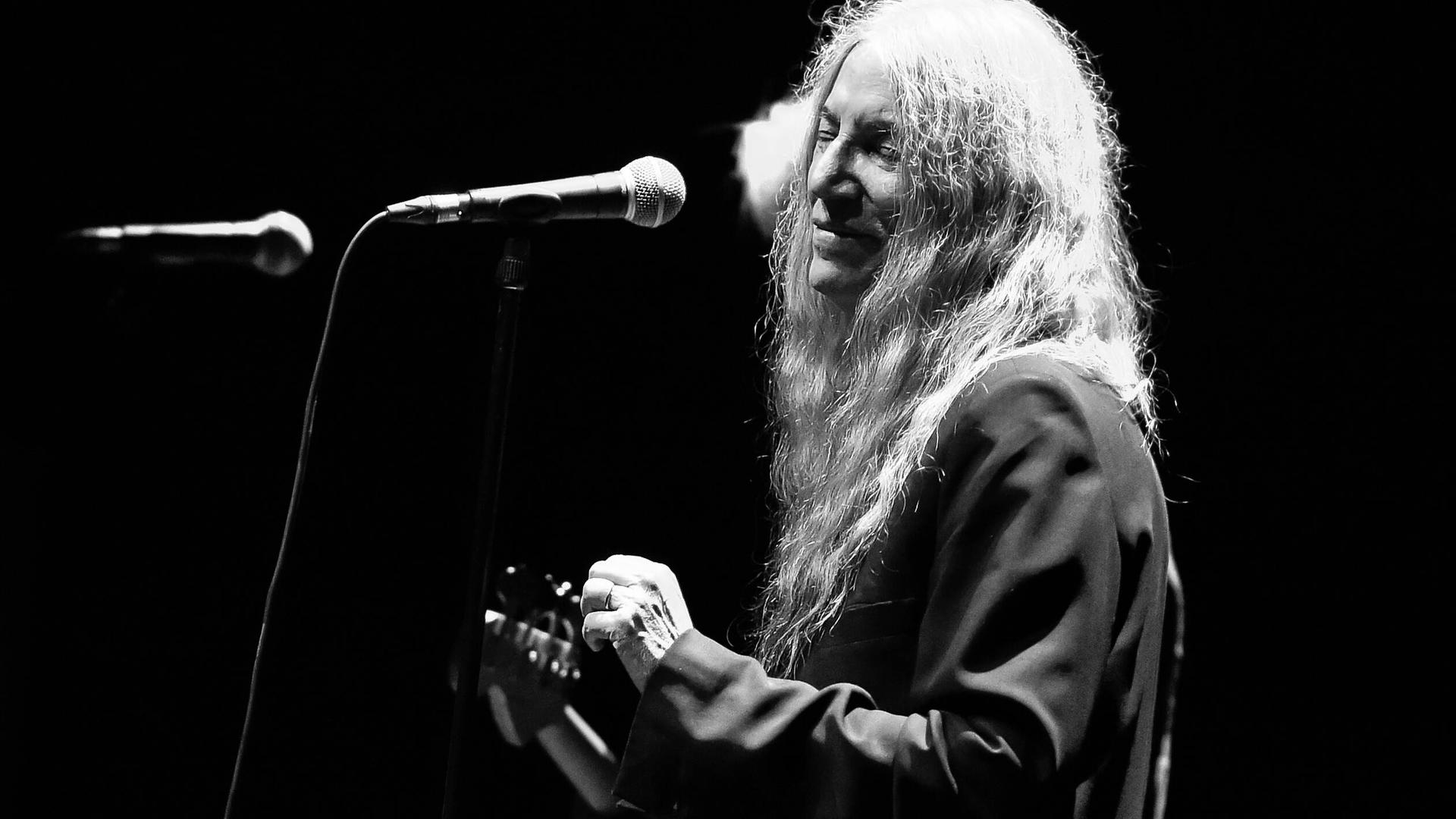 Schwarzweiß Aufnahme von Patti Smith auf der Bühne. Sie hat die Gitarre in der Hand und steht vor einem Mikrofon. Schwarzweiß Aufnahme von Patti Smith auf der Bühne. Sie hat die Gitarre in der Hand und steht vor einem Mikrofon.
