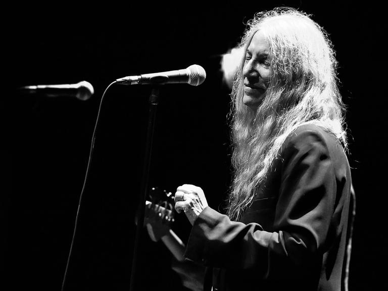 Schwarzweiß Aufnahme von Patti Smith auf der Bühne. Sie hat die Gitarre in der Hand und steht vor einem Mikrofon. 