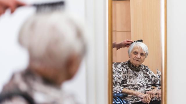Eine alte Frau im Rollstuhl sitzt vor einem Spiegel. Sie bekommt die Haare von einer Altenpflegerin gekämmt.