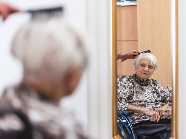 Eine alte Frau im Rollstuhl sitzt vor einem Spiegel. Sie bekommt die Haare von einer Altenpflegerin gekämmt.