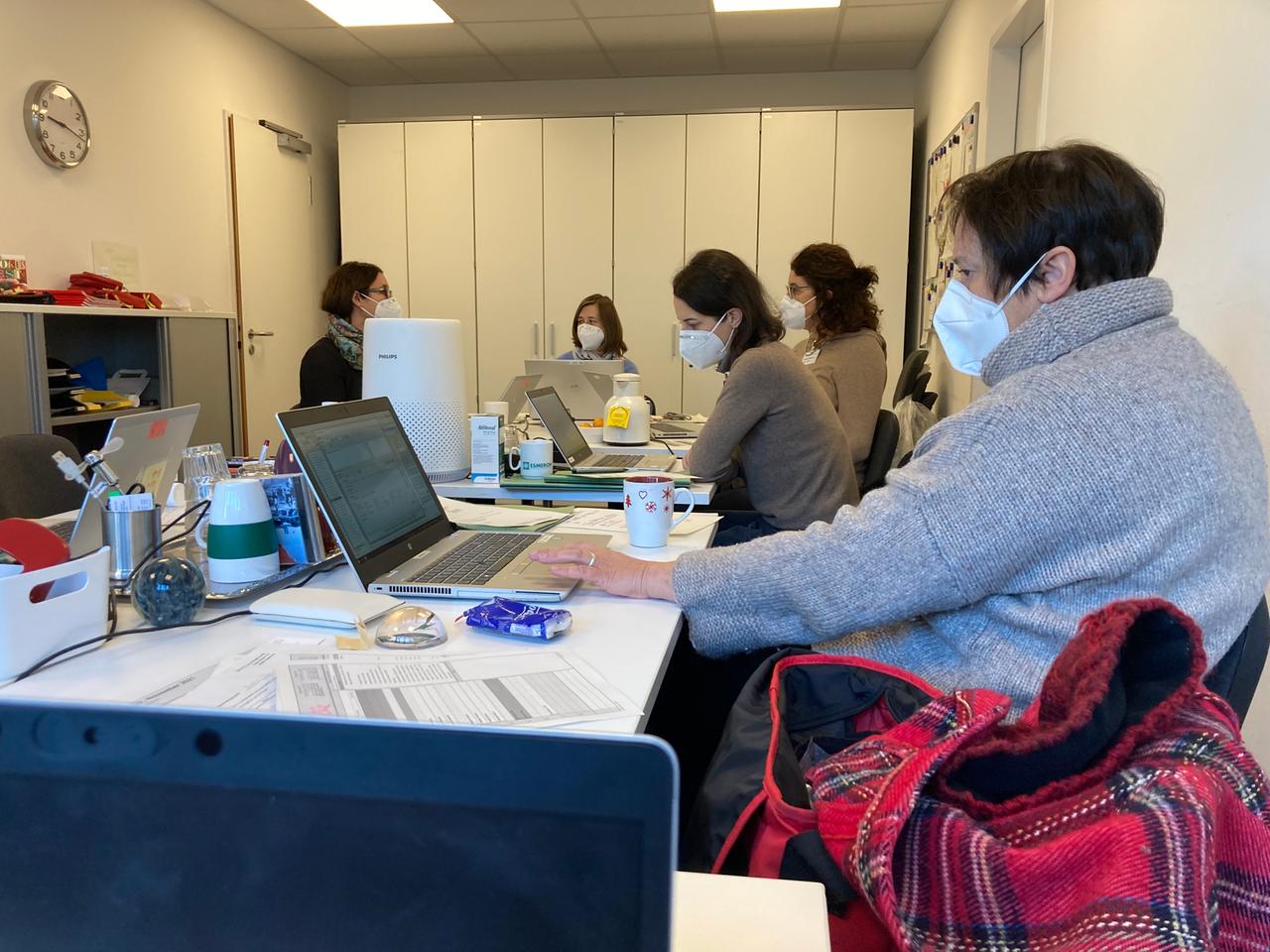Fünf Menschen sitzen mit FFP-2-Masken in einem Raum vor ihren Laptops. Fünf Menschen sitzen mit FFP-2-Masken in einem Raum vor ihren Laptops.