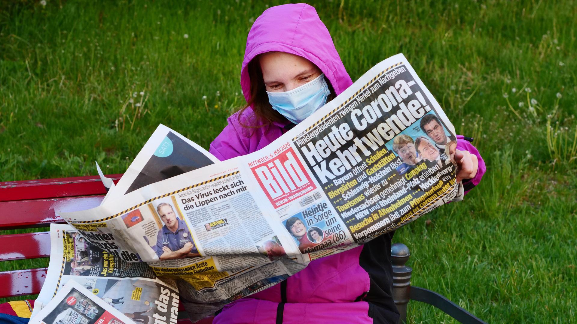 Eine Frauen traegt eine Schutzmaske zum Schutz gegen das neuartige Coronavirus, waehrend sie auf einer Bank sitzt und die Bildzeitung liest. Sie hatte die Zeitung wegen der Informationen zu den neuesten Corona-Regeln gekauft. Die Titelschlagzeile lautet: "Heute Corona-Kehrtwende!". Eine Frauen traegt eine Schutzmaske zum Schutz gegen das neuartige Coronavirus, waehrend sie auf einer Bank sitzt und die Bildzeitung liest. Sie hatte die Zeitung wegen der Informationen zu den neuesten Corona-Regeln gekauft. Die Titelschlagzeile lautet: "Heute Corona-Kehrtwende!".