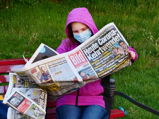 Eine Frauen traegt eine Schutzmaske zum Schutz gegen das neuartige Coronavirus, waehrend sie auf einer Bank sitzt und die Bildzeitung liest. Sie hatte die Zeitung wegen der Informationen zu den neuesten Corona-Regeln gekauft. Die Titelschlagzeile lautet: "Heute Corona-Kehrtwende!". 