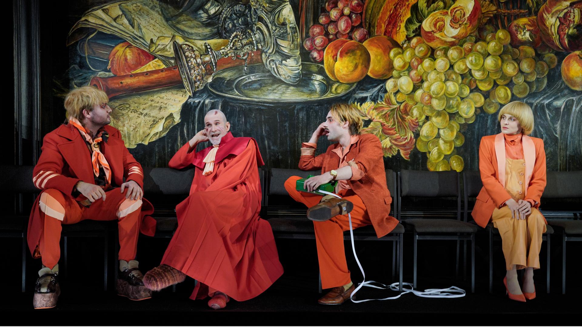 Szenefoto: Vier Schauspielerinnen und Schauspieler sitzen vor einem überdimensionierten Stillleben-Gemälde. Szenefoto: Vier Schauspielerinnen und Schauspieler sitzen vor einem überdimensionierten Stillleben-Gemälde.