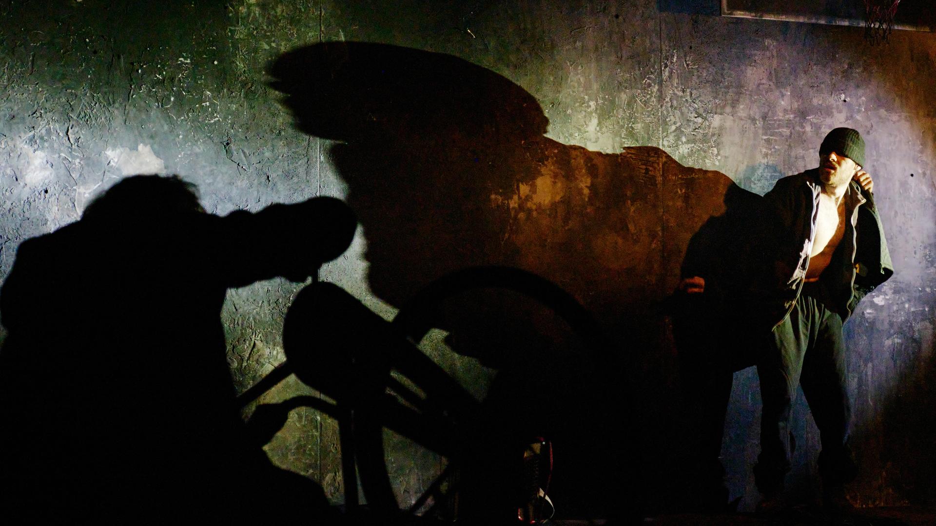Ein Schauspieler mit tief ins Gesicht gezogener Mütze wirft einen langen Schatten an die Wand.