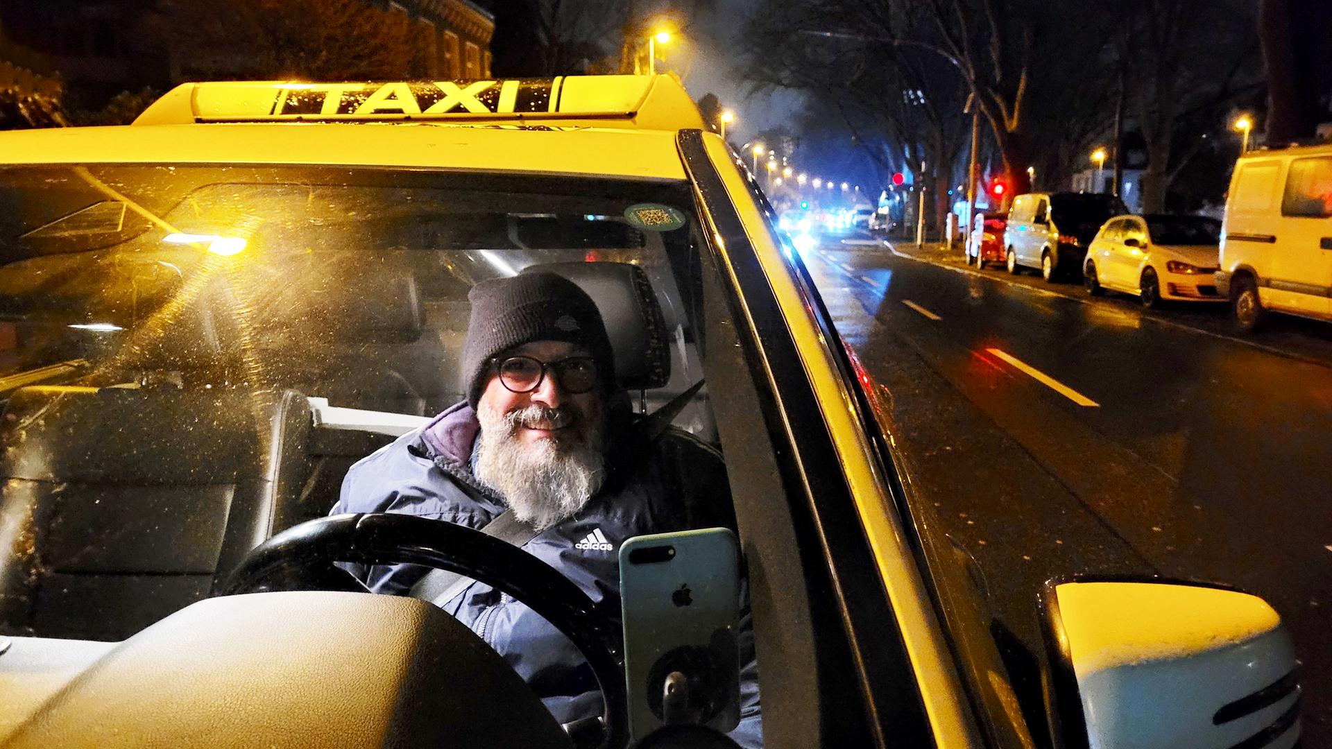 Auf einer Straße steht ein Taxi, in der ein bärtiger Taxifahrer sitzt, der eine Brille trägt. 