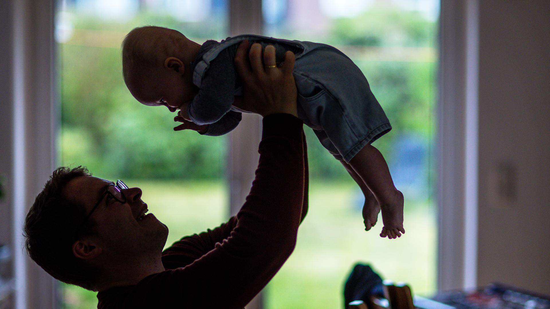 Ein junger Vater spielt mit seinem sechs Monate alten Sohn im Wohnzimmer, hält ihn in die Luft, der Säugling lacht Ein junger Vater spielt mit seinem sechs Monate alten Sohn im Wohnzimmer, hält ihn in die Luft, der Säugling lacht