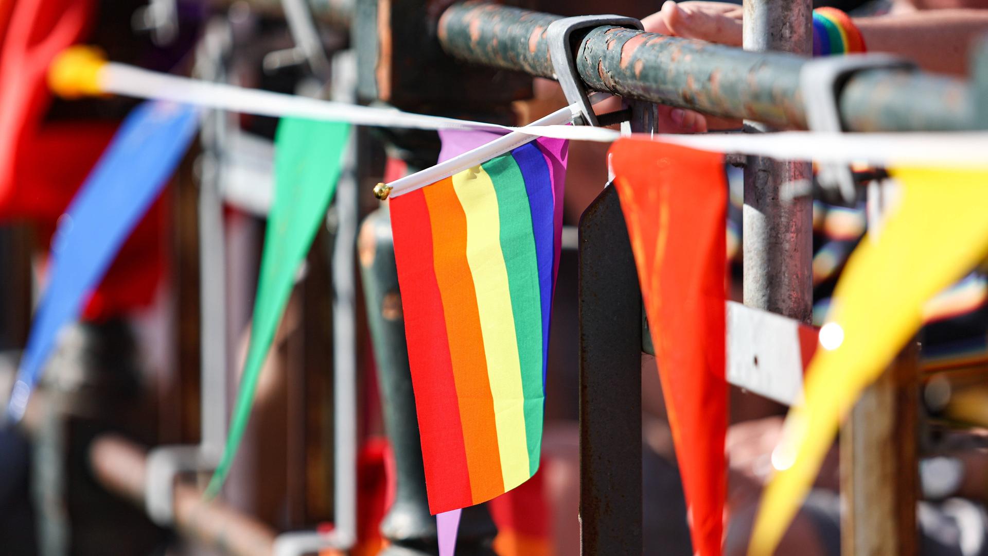 An einem Brückengeländer sind verschiedene Bänder und Fahnen in bunten Farben befestigt. Im Bildzentrum: Die Regenbogenfahne der LGBTQ-Community. An einem Brückengeländer sind verschiedene Bänder und Fahnen in bunten Farben befestigt. Im Bildzentrum: Die Regenbogenfahne der LGBTQ-Community.