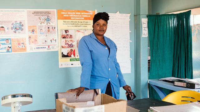 Die Gesundheitsassistentin Priscar Mailosi steht in einer Klinik, in blau gekleidet mit dunklem Stirnband im Haar. Im Hintergrund Plakate zur Gesundheitsvorsorge.