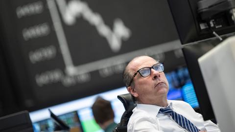 Ein Mann mit Brille schaut auf seinen Monitor, im Hintergrund eine Anzeigetafel mit dem Kursverlauf an der Börse.