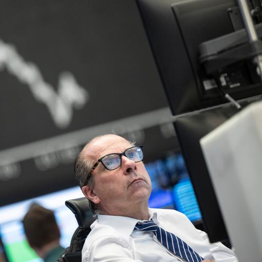 Ein Mann mit Brille schaut auf seinen Monitor, im Hintergrund eine Anzeigetafel mit dem Kursverlauf an der Börse.