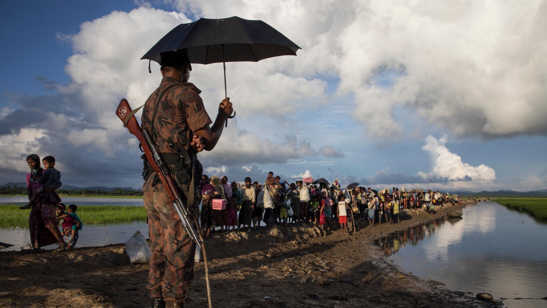 Ein Wächter aus Bangladesch mit geschultertem Gewehr steht unter einem Sonnenschirm während Tausende Rohingya-Flüchtlinge aus Myanmar an einem schlammigen Reisfeld entlanggehen.
