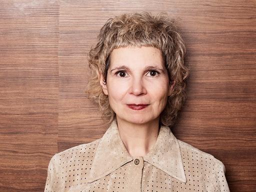 Eine Frau mit blonden kurzen Haaren steht vor einer dunkelbraunen Holzwand. Sie blickt freundlich in die Kamera und trägt eine braune Bluse aus einem festen Stoff.