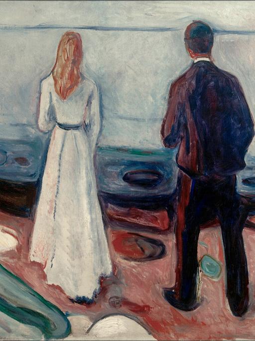 Ein Paar steht am Strand und schaut auf's Meer. Der Mann steht ein wenig hinter der Frau und scheint ebenfalls auf sie zu blicken.