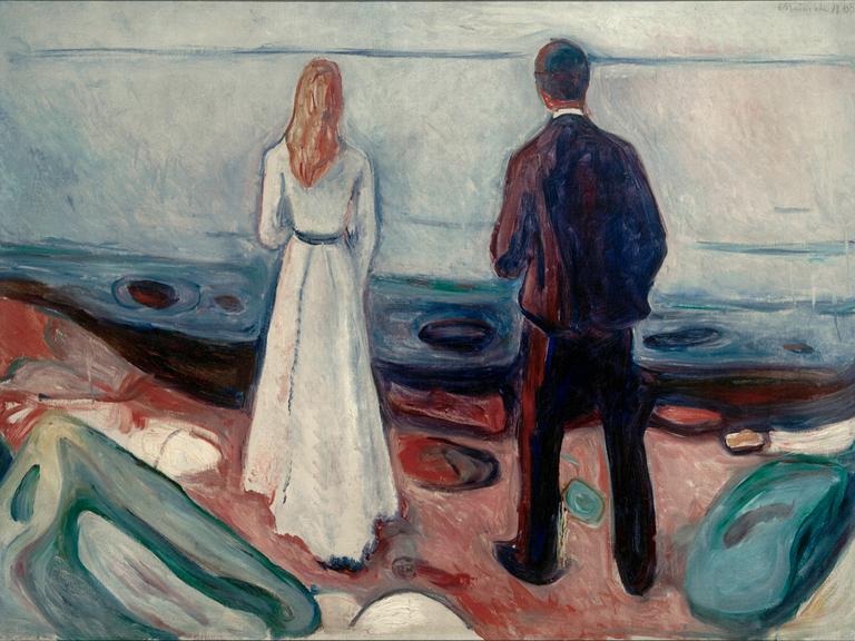 Ein Paar steht am Strand und schaut auf's Meer. Der Mann steht ein wenig hinter der Frau und scheint ebenfalls auf sie zu blicken.