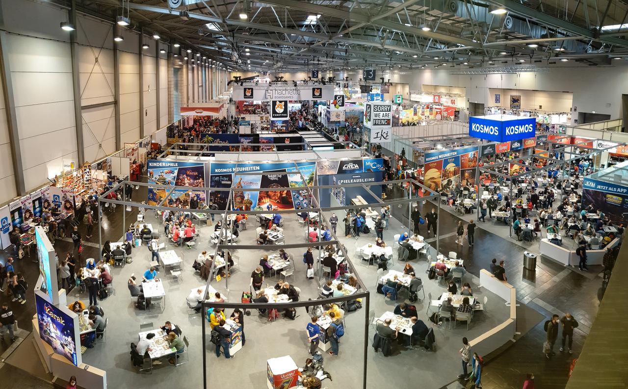 Eine Messehalle von oben fotografiert: Es befinden sich mehrere Tische im Raum, an denen verschiedene nicht-elektronische Spiele gespielt werden. Eine Messehalle von oben fotografiert: Es befinden sich mehrere Tische im Raum, an denen verschiedene nicht-elektronische Spiele gespielt werden.