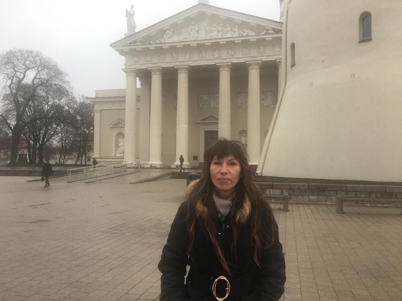 Eine Frau im Wintermantel steht auf einem Platz, im Hintergrund ein weißes Gebäude mit Säulen: Elena Kotenochkina Eine Frau im Wintermantel steht auf einem Platz, im Hintergrund ein weißes Gebäude mit Säulen: Elena Kotenochkina