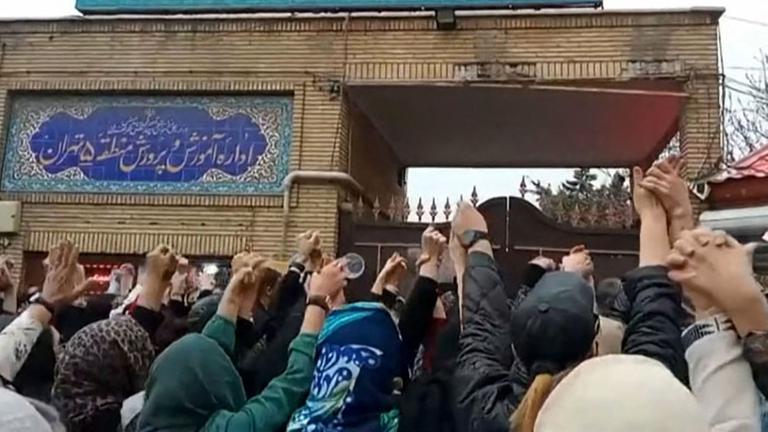 Viele Menschen stehen vor einem Gebäude. Die meisten heben ihre Arme in die Luft. Das Gebäude ist das Bildungs-Ministerium in dem Land Iran.