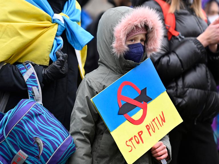 Ein junger Demonstrant hält ein Plakat in den Farben der ukrainischen Flagge, mit der Aufschrift „Stop Putin“ und der Darstellung einer Rakete, die mit einem roten Verbotszeichen versehen ist, in seinen Händen.