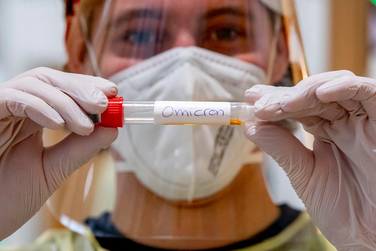 Ein Labormitarbeiter hält vor sich ein Teströhrchen mit der Aufschrift "Omicron". Er trägt Handschuhe und eine Maske vor Mund und Nase.