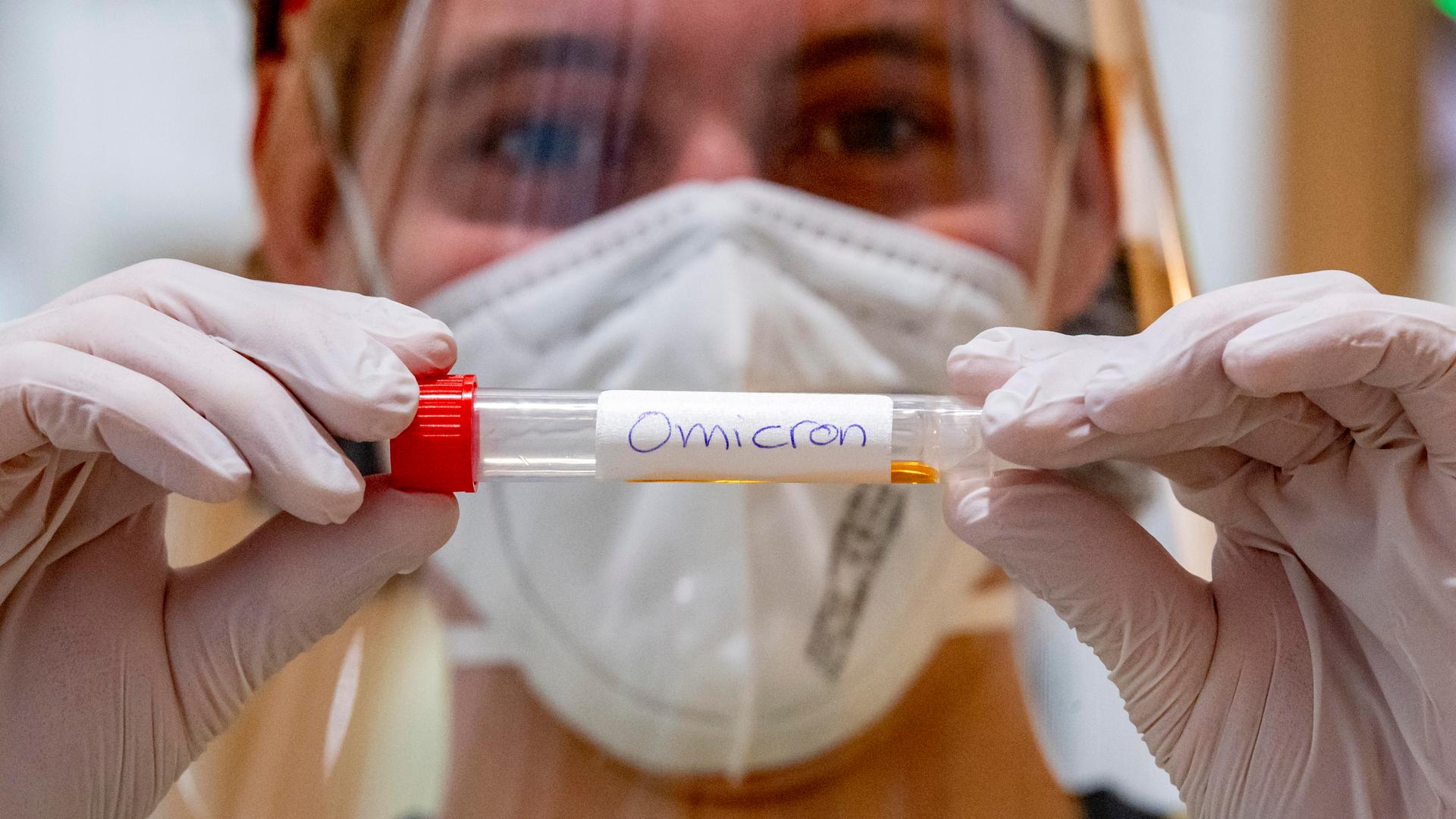 Ein Labormitarbeiter hält vor sich ein Teströhrchen mit der Aufschrift "Omicron". Er trägt Handschuhe und eine Maske vor Mund und Nase.
