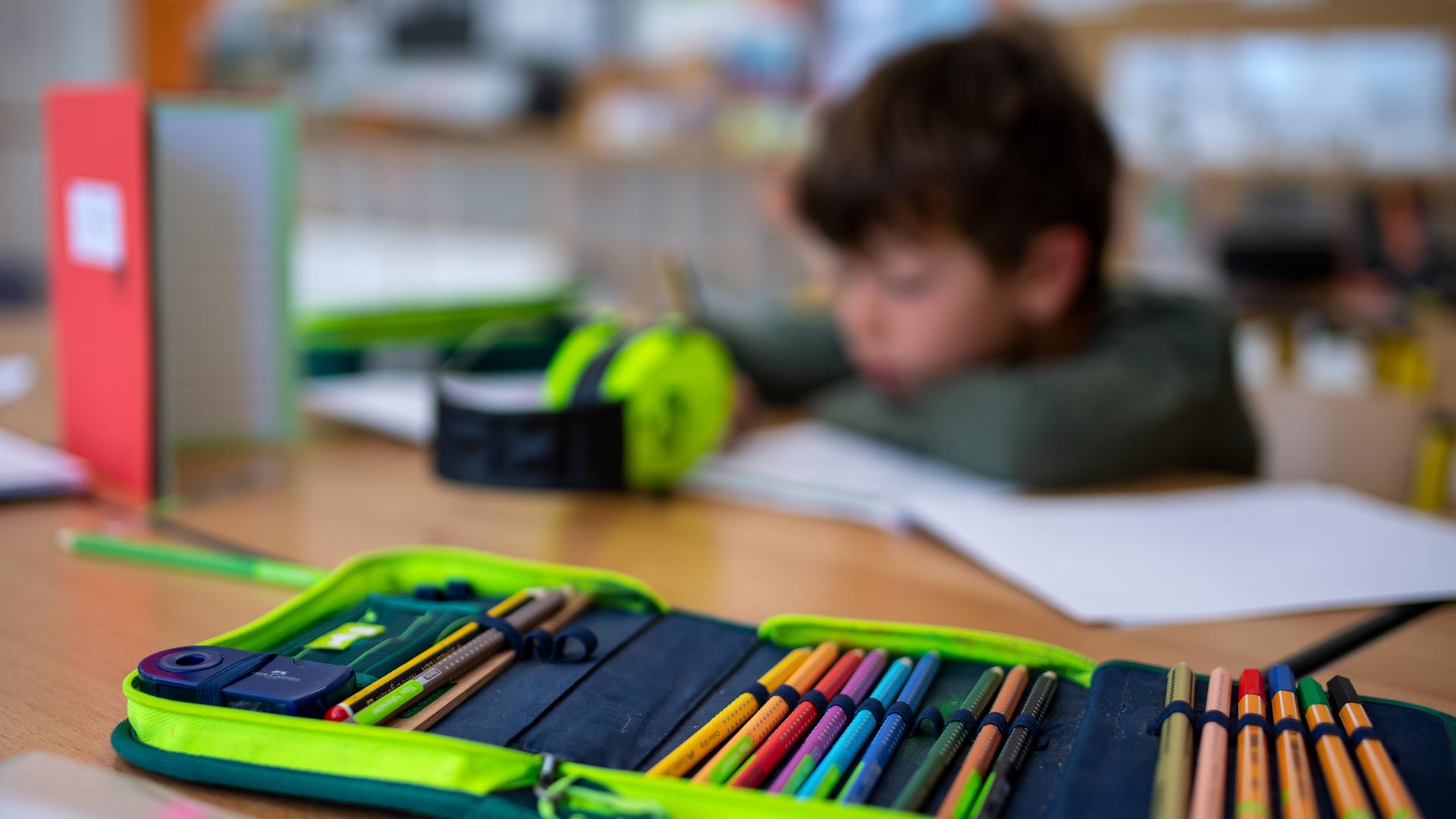Ein Junge in einer Schulklasse sitzt an einem Tisch und schreibt, im Vordergrund ein Mäppchen mit Stiften.