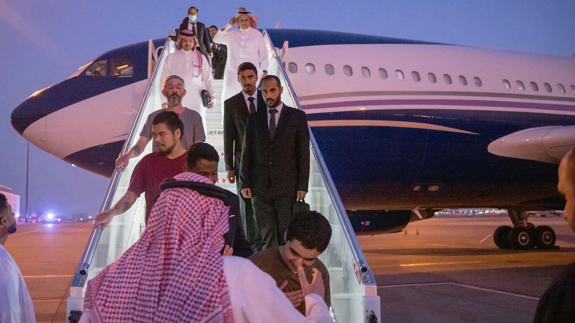 Mehrere Männer, teils in Anzügen, teils leger gekleidet, kommen die Treppe aus einem Flugzeug runter. Unten steht ein Mann mit traditioneller saudi-arabischer Kleidung und Kopftuch. Man sieht ihn von hinten.