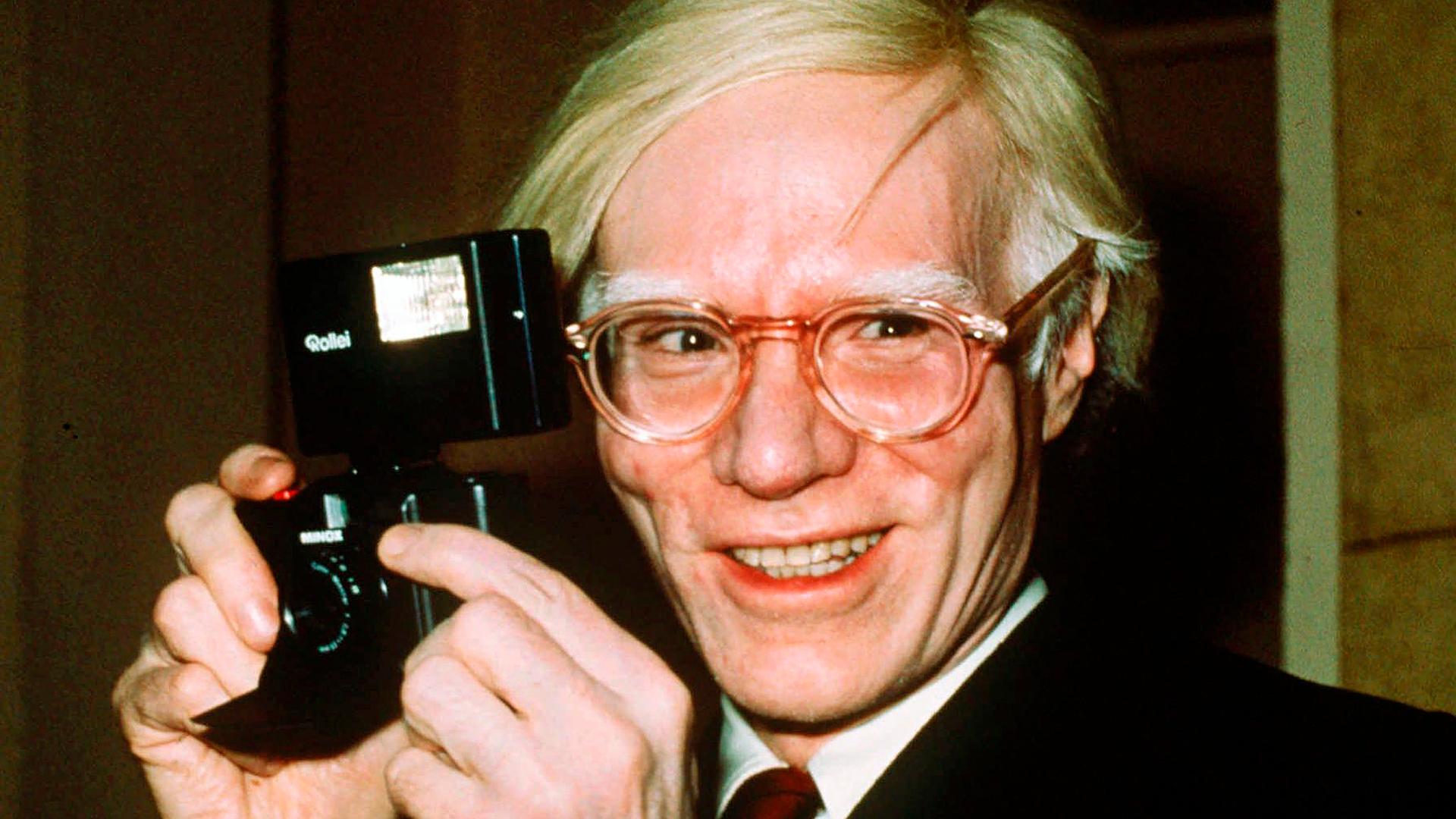 Der Künstler Andy Warhol mit einer Kamera in den Händen. 