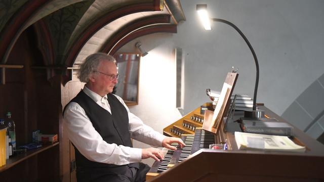 Ein Mann sitzt vor dem Spieltisch einer Orgel, die Hände sind auf den Tasten. Er trägt weißes Hemd und schwarze Weste. Er blickt konzentriert auf die Noten vor sich.