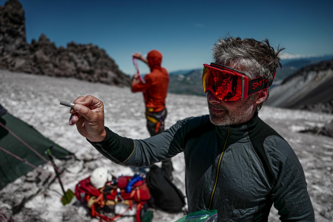 Ein Mann mit Skibrille steht im Schnee und hält ein kleinen Metallstift der Kamera entgegen.