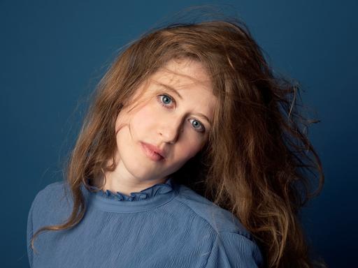 Wir sehen eine Frau, die einen blauen Pullover trägt und in die Kamera guckt. 