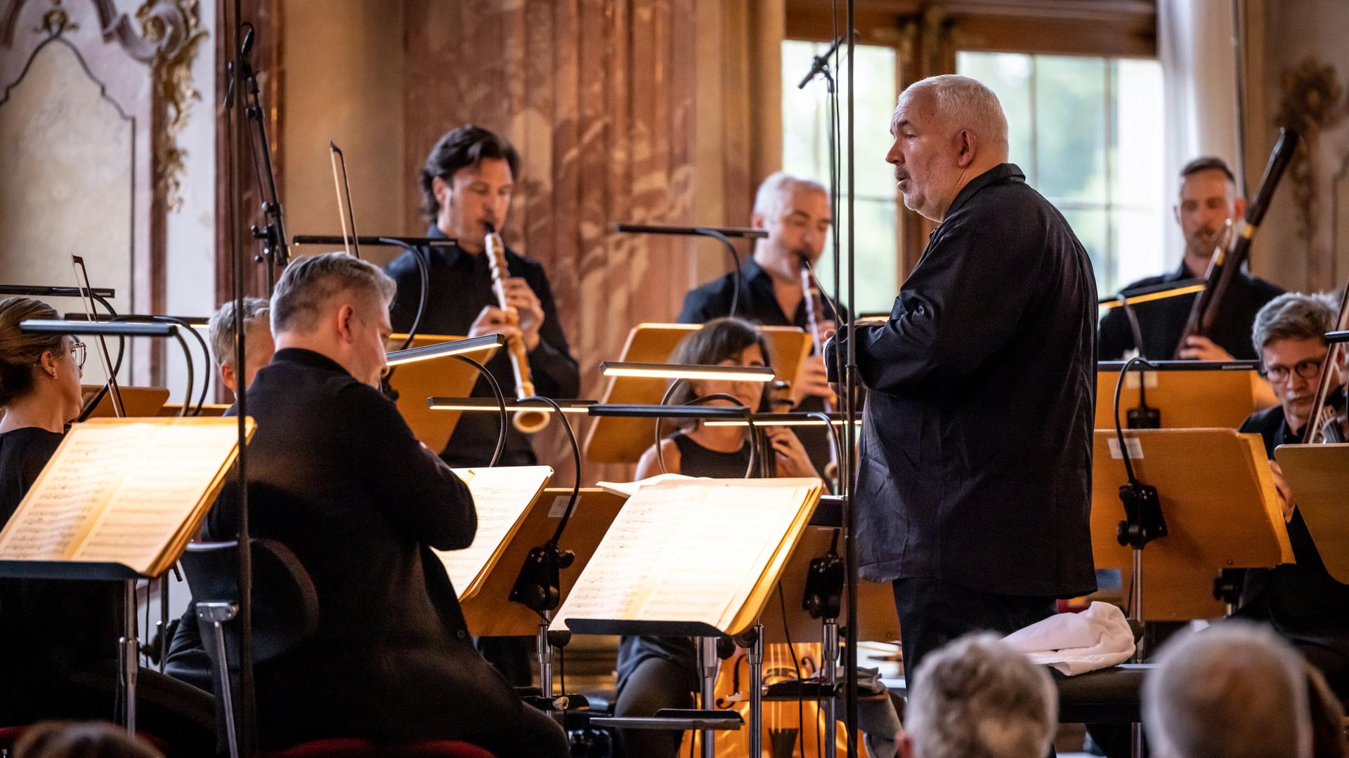 Ein Orchester ist zu sehen und der Dirigent der am Pult vor dem Orchester steht. Ein Orchester ist zu sehen und der Dirigent der am Pult vor dem Orchester steht.