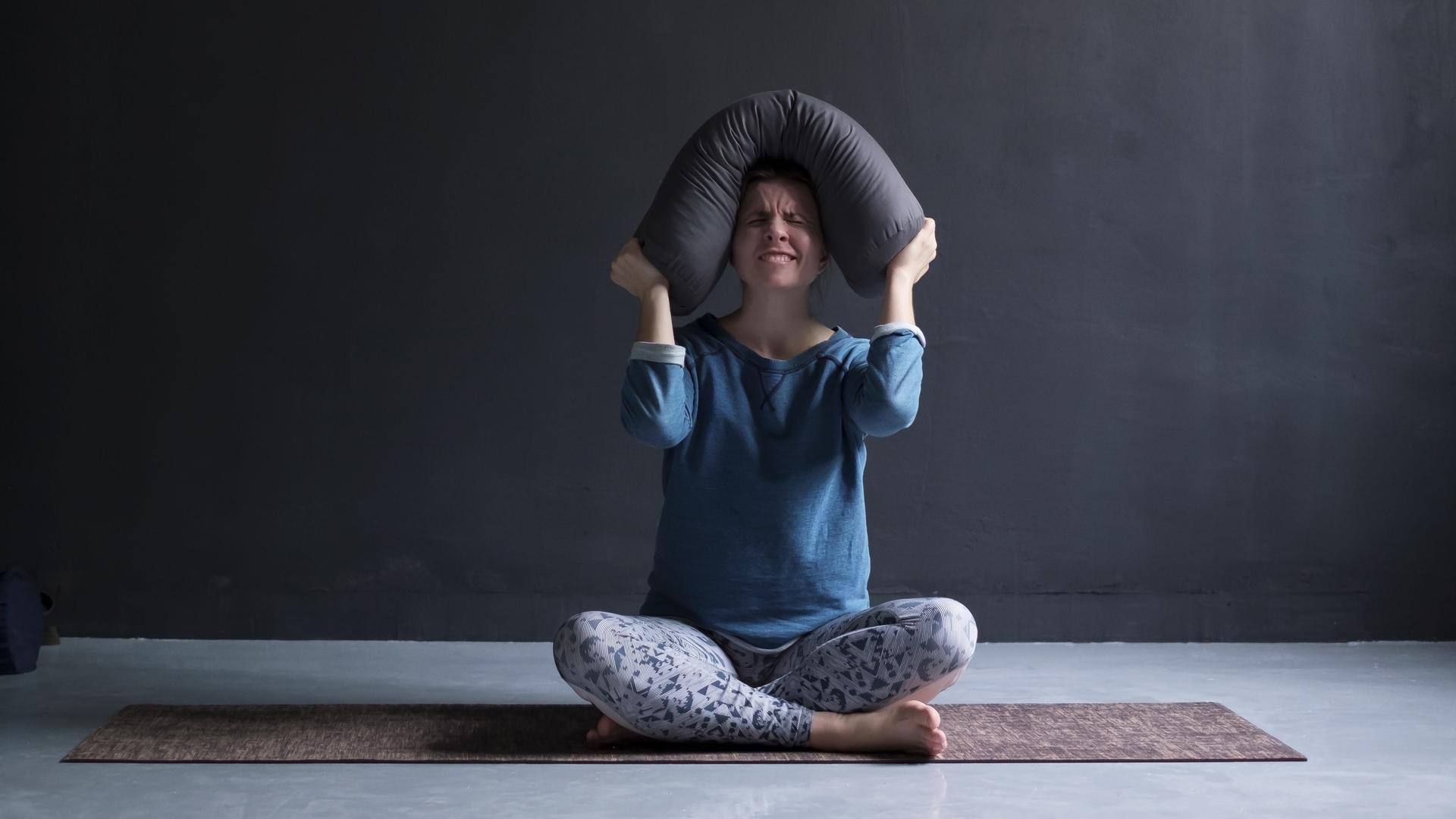 Eine Frau sitzt auf einer Yogamatte und drückt wütend ein Kissen auf ihren Kopf.