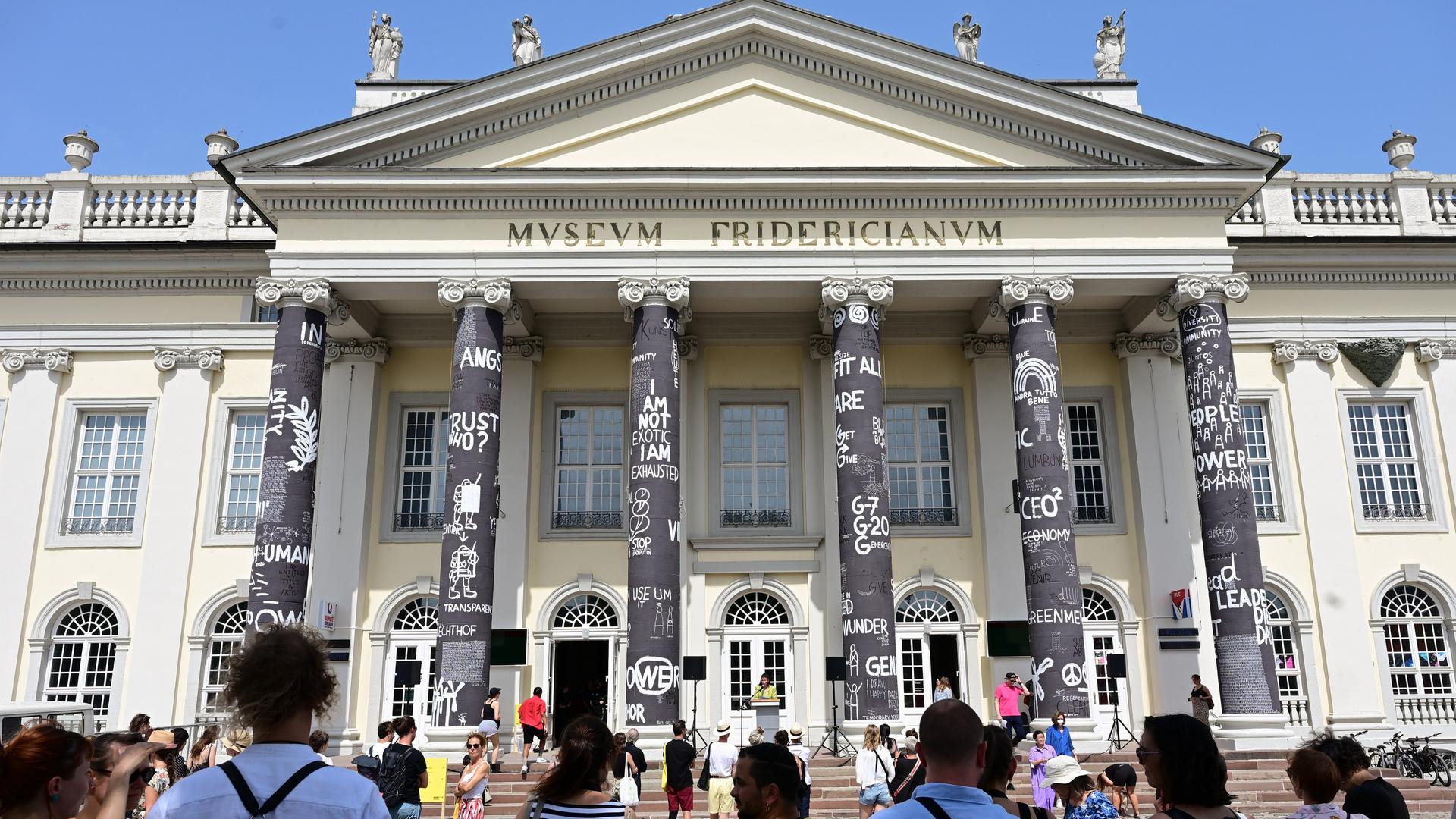 Besucher der documenta 15 stehen nach Eröffnung vor dem Fridericianum, dessen Säulen der rumänische Künstler Dan Perjovschi mit schwarzer Farbe bemalt und mit weißen Symbolen und Zeichen zu Themen wie Frieden, Solidarität oder Nachhaltigkeit beschriftet hat