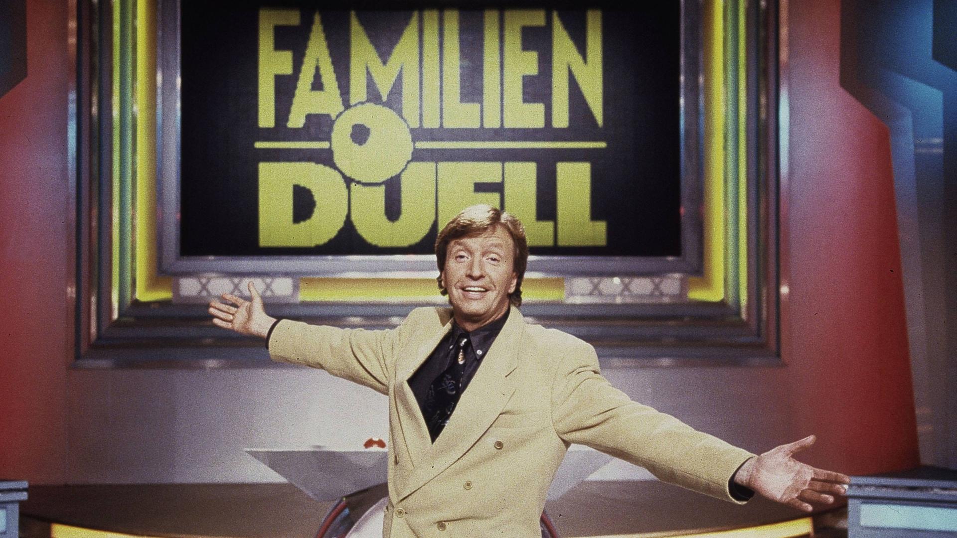 Moderator Werner Schulze-Erdel in einem beigen Sakko mit einladend ausgebreiteten Armen in der Kulisse der RTL-Quizshow "Familienduell", deren Titel hinter ihm in goldenen Lettern auf einem großen Bildschirm steht. Moderator Werner Schulze-Erdel in einem beigen Sakko mit einladend ausgebreiteten Armen in der Kulisse der RTL-Quizshow "Familienduell", deren Titel hinter ihm in goldenen Lettern auf einem großen Bildschirm steht.