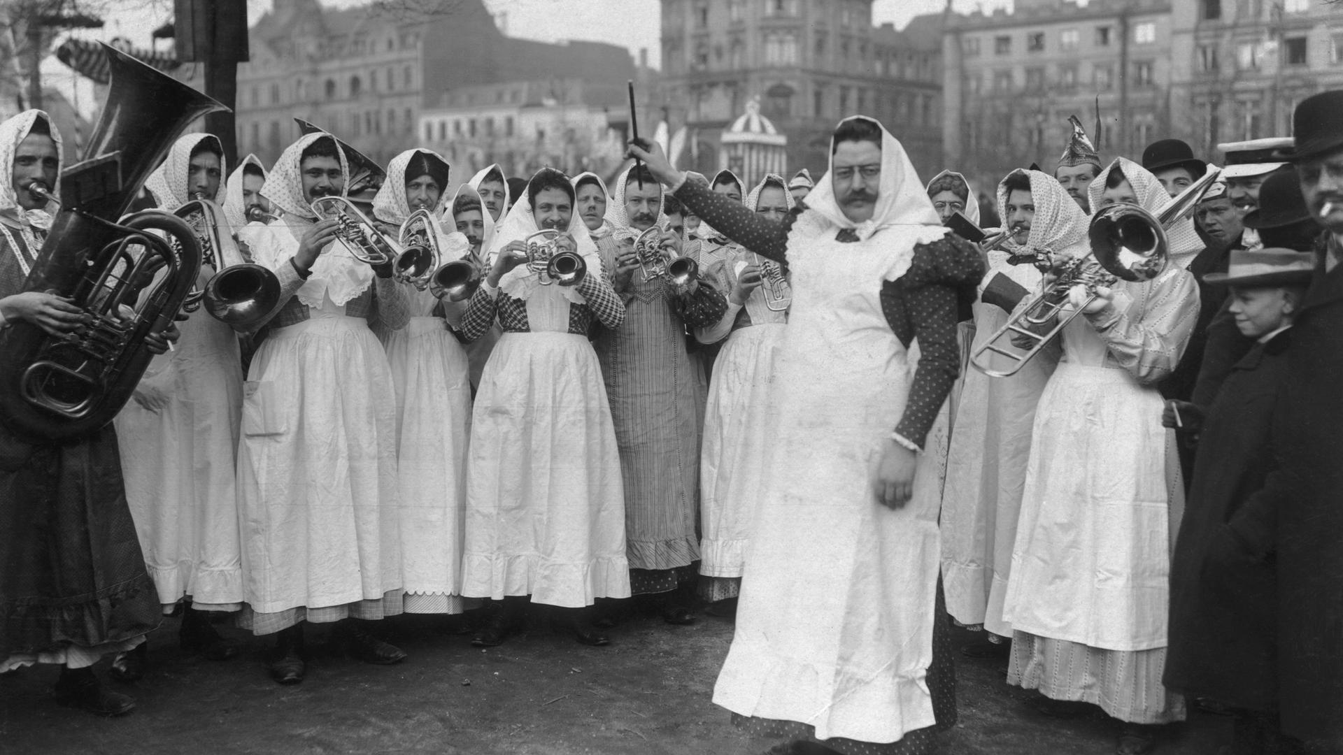 Das Orchester der "Kölner Milchmädchen" tritt in Verkleidung beim Kölner Strassenkarneval auf, ca. 1910. Eine Männerguppe in Frauenkleidung mit Kopftüchern spielt mit Blasinstrumenten einen Tusch.